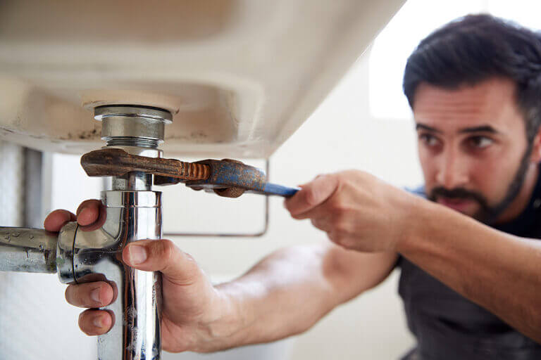 Male Plumber Using Wrench To Fix Leaking Sink In Home Bathroom